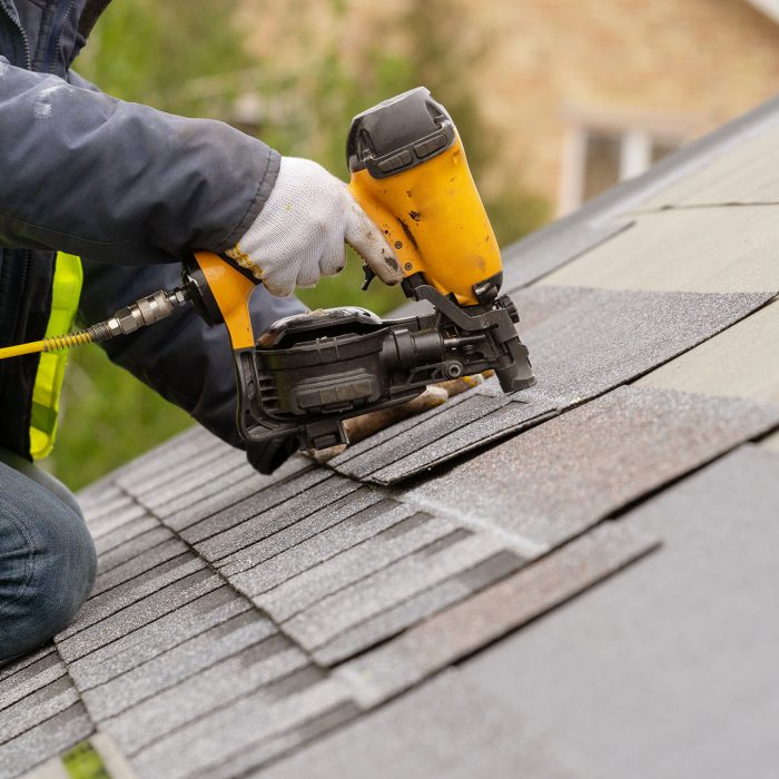Roofer worker in special protective work wear and gloves, using air or pneumatic nail gun and installing asphalt or bitumen shingle on top of the new roof under construction residential building